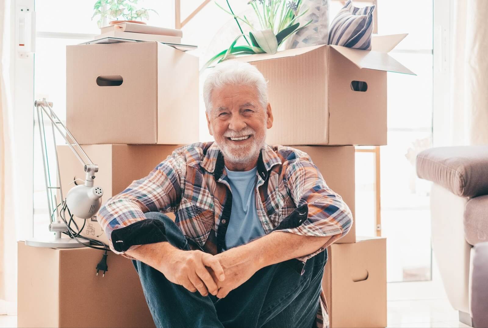 A senior man smiling while sitting on the floor in front of moving boxes.