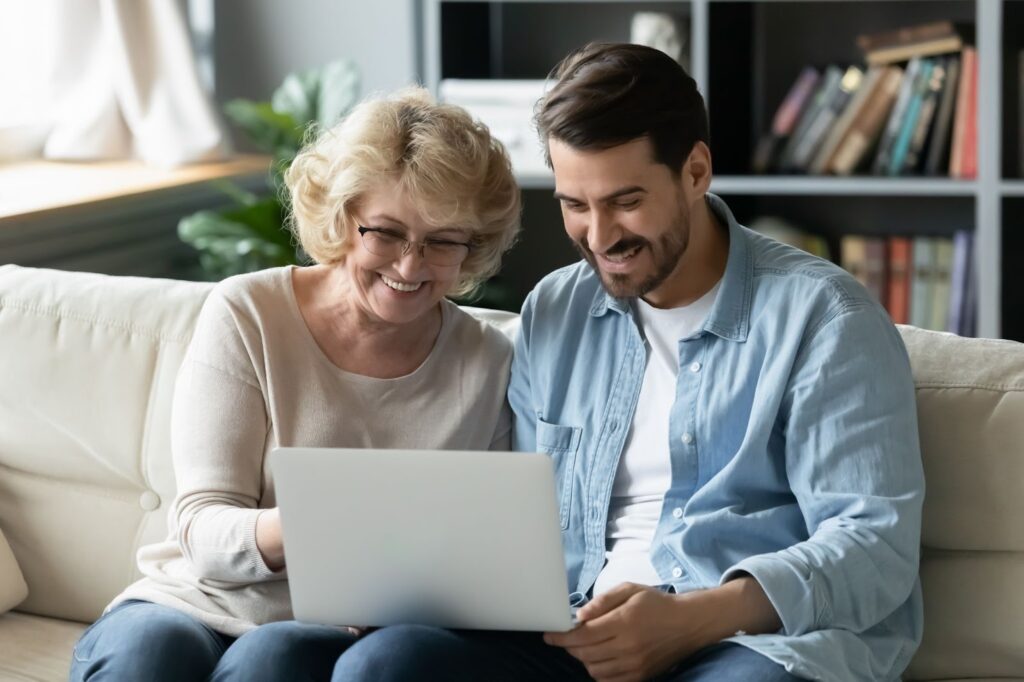 A young man and his older mother smile while using a computer to talk about his mother moving to assisted living.