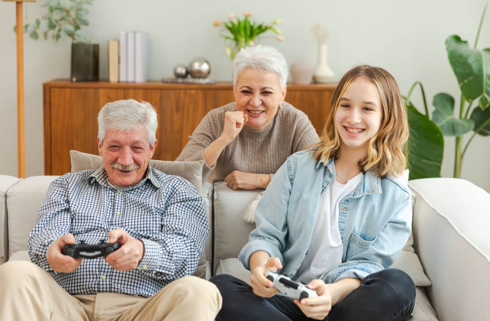 A senior holds a video game controller, playing on the couch with their grandchild and their partner rooting behind them