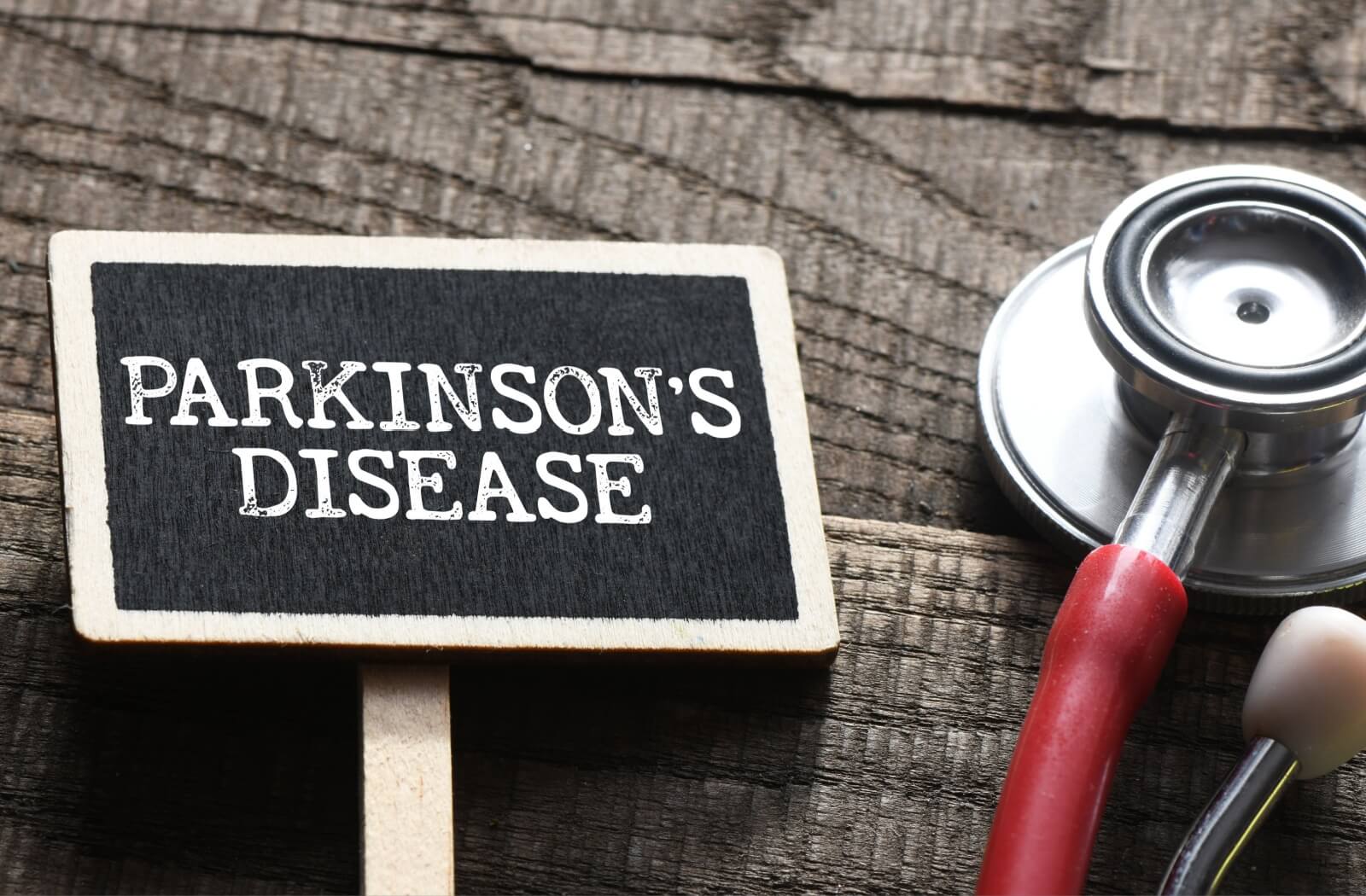 A black sign with the words "Parkinson’s Disease" written in white sits on a wooden surface, next to a red stethoscope.