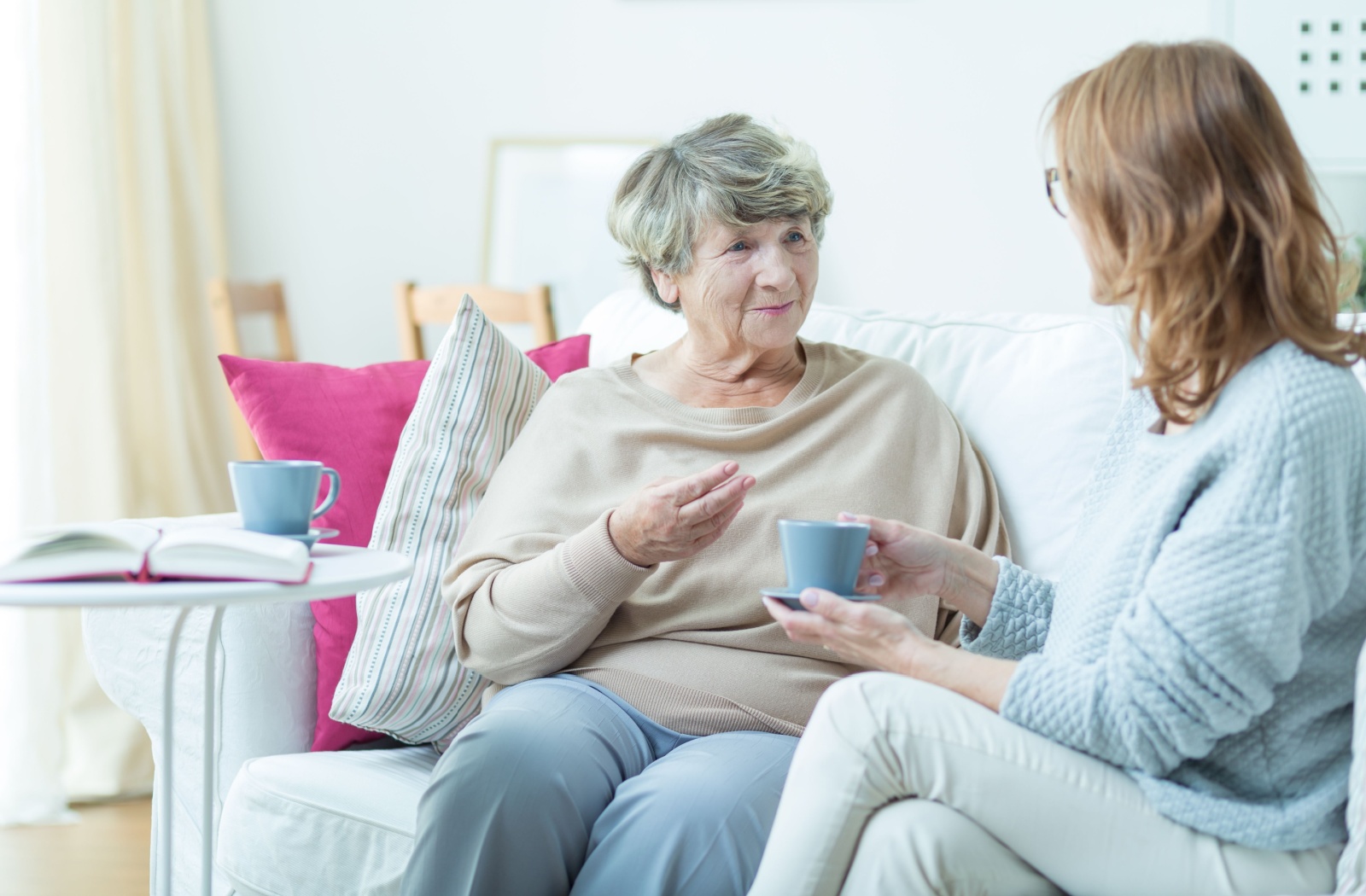 Senior woman and adult daughter talk to each other on the couch while drinking from blue mugs.