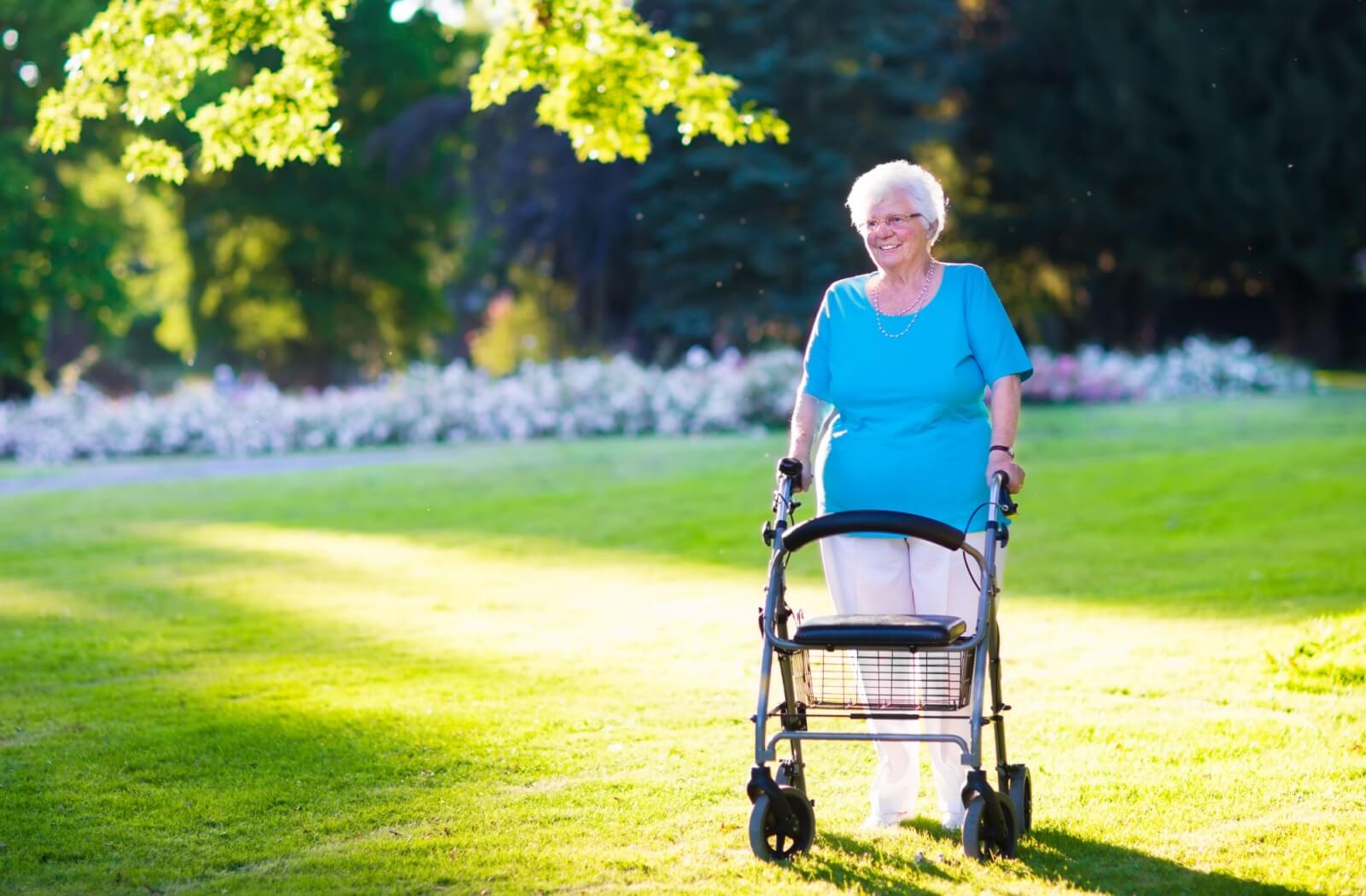 Person using a walker smiles while walking through a sunlit park with green grass and trees in the background