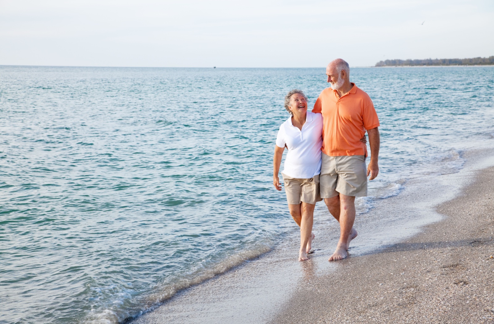 A laughing retired couple walks across the shore of a Florida beach in the afternoon with their arms around one another