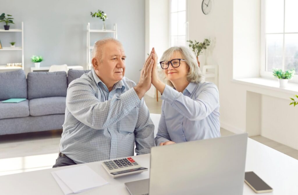 A couple sitting at a desk in a well-lit room high-fives after planning out their budgets. In front of them, they have a calculator and a laptop for planning purposes.