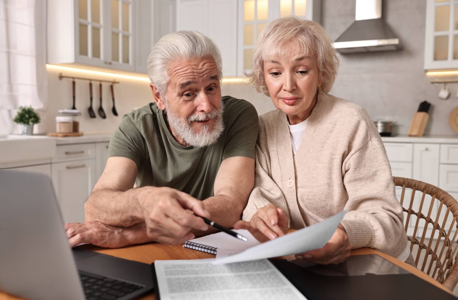 An older couple sits in their kitchen at their laptop, with papers in front of them. They peer down at the paper, pointing to it with a pen while they work on their budget.