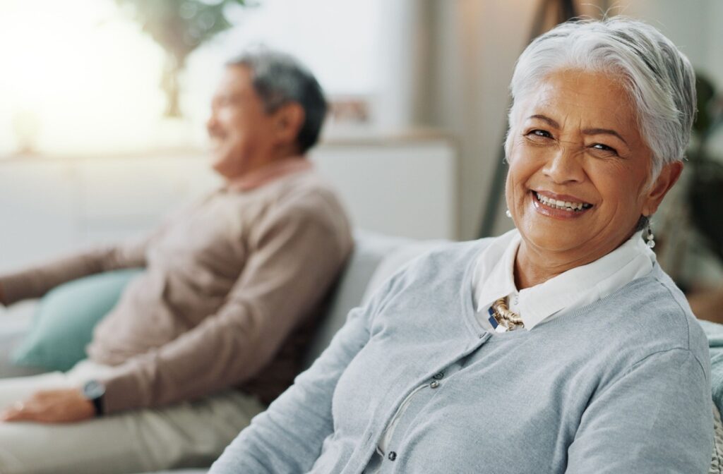  A senior smiles at the camera while their partner sits on the couch with them.
